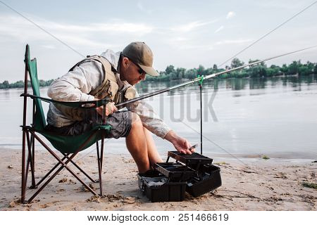 Fisherman Is Sitting In Folding Chair And Leaning Forward To Opened Black Plastic Box. Guy Holds Fly