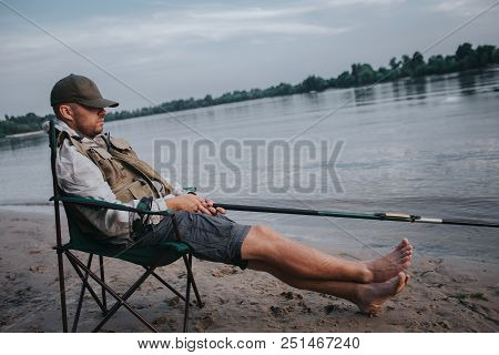 Fisherman Is Sitting In Folding Chair And Sleeping. His Cap Is Lying On Eyes. Man Holds Fly Rod In H