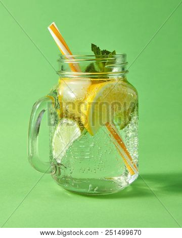 Glass mason jar on a green table with homemade sparkling lemonade with ice, slices of lime and lemon, green leaves of mint with plastic straws.