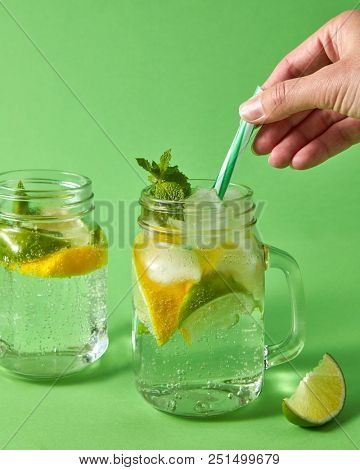 Citrus fruits slices of lemon and lime, ice, leaf of mint, water. Two glass jar on green background with plastic straws with famale hand. Cold natural lemonade handmade with bubbles of air.