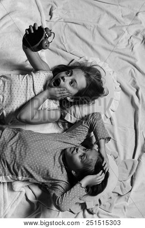 Girls Lie On White And Pink Bed Sheets Holding Red Alarm Clock. Morning And Deadline Concept. School