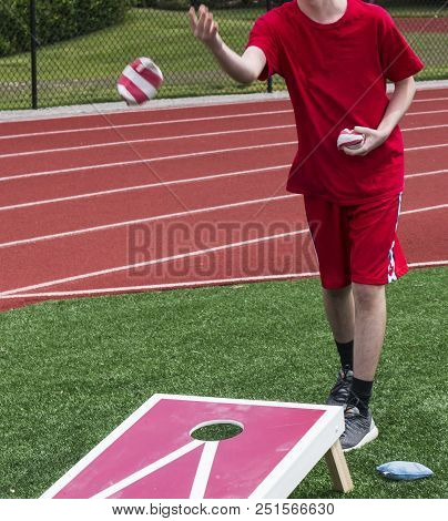 A Teenage Boy Tosses A Bean Bag While Playing Corn Hole In Gym Class On The Turf With The Track Behi