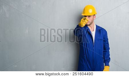 Young caucasian man over grey grunge wall wearing contractor uniform and safety helmet pointing with finger to the camera and to you, hand sign, positive and confident gesture from the front