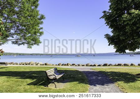 Bench In The Park At Canandaigua Lake. View Of The Calm Blue Lake, Sidewalk To The Lakeshore In The 