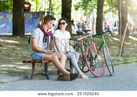 Young Couple In Love With Bicycle Sitting On Beanch In The Sunny Summer Park