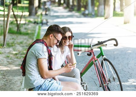 Young Romantic Couple With Bicycle Sitting On Beanch And Speaking In The Sunny Summer Park