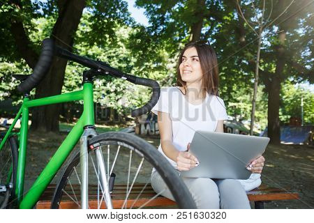 Cute Romantic Girl With Green Bicycle Sitting On Beanch And Working With Laptop In The Sunny Summer 