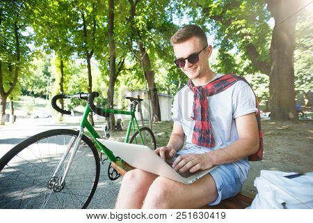 Handsome Hipster Boy In Sunglasses With Green Bicycle Sitting On Beanch And Working With Laptop In T
