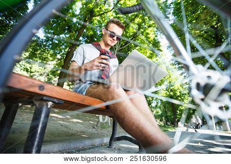 Handsome Hipster Boy In Sunglasses With Green Bicycle Sitting On Beanch And Working With Laptop In T