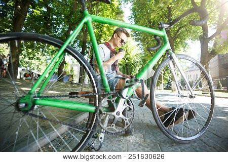 Handsome Hipster Boy In Sunglasses With Green Bicycle Sitting On Beanch And Working With Laptop In T