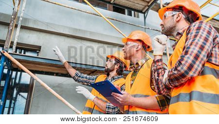 Wide Shot Of Group Of Builders In Hard Hats And Reflective Vests Pointing At Building House