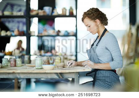 Young creative woman in workwear sitting by table in clay craft workshop and preparing small details for earthenware items