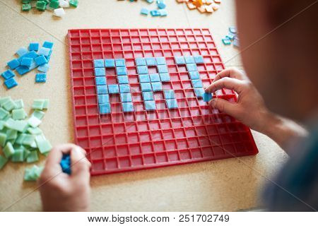 Over the shoulder view of unrecognizable craftsman laying out  word ART in blue square tiles over red base, copy space