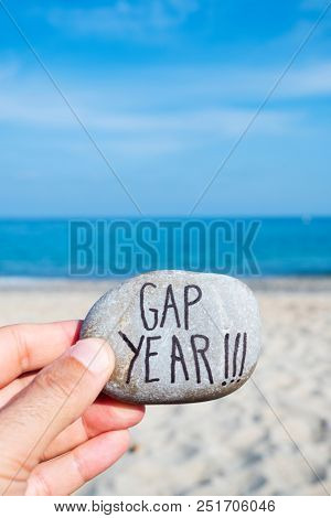 closeup of the hand of a young caucasian man on the beach, in front of the ocean, holding a stone with the text gap year written in it