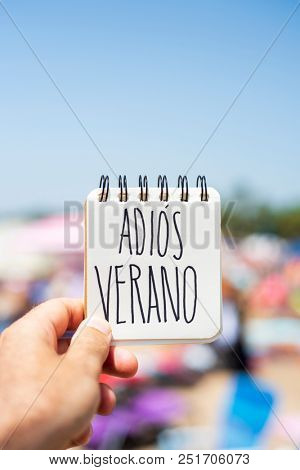 closeup of the hand of a young caucasian man in the seashore of a beach showing a spiral notepad with the text adios verano, good bye summer in spanish, handwritten in it