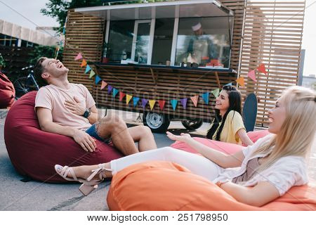 Happy Young Friends Laughing While Sitting On Bean Bag Chairs Near Food Truck