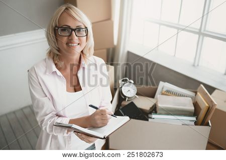 Smiling Blond Woman With Glasses With Notebook Makes List Of Things In Cardboard Box While Moving To