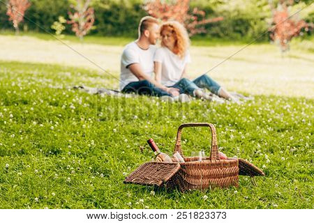 Close-up View Of Picnic Basket With Bottle Of Wine And Couple Sitting On Lawn Bhind At Park