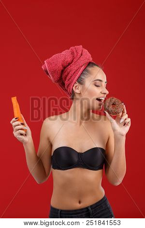 A Smiling Girl Is Eating A Chocolate Donut, Prefering It To A Healthy Carrot, Standing On The Red Ba