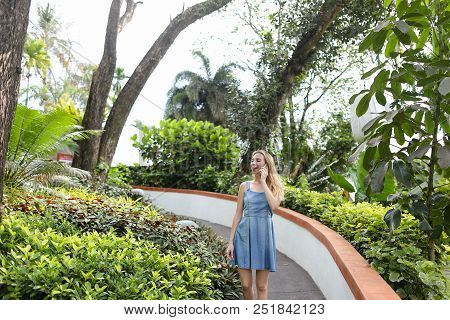 Young Woman Talking By Smartphone Near Tropical Plants, Wearing Jeans Sundress.