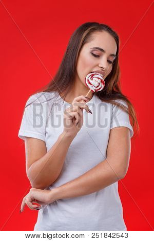 A Positive Girl With Dark Long Hair Is Licking A Sugar Candy, Posing On The Red Background.