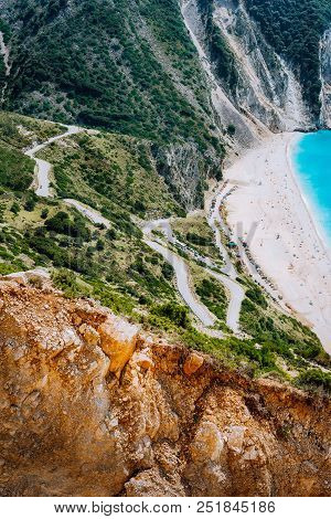 Serpentine Zigzag Road Down To Famous Myrtos Beach. Orange Rocks Ravine On Side. Sunny Beach From Vi