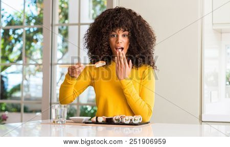 African american woman eating sushi using chopsticks at home cover mouth with hand shocked with shame for mistake, expression of fear, scared in silence, secret concept