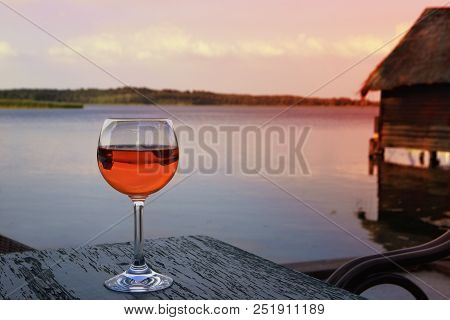 Glass With Spritz Veneziano On A Wooden Table Overlooking The Lake With A Boathouse At Sunset, A Fam