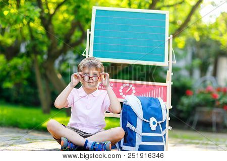 Happy Little Kid Boy With Glasses Sitting By Desk And Backpack Or Satchel. Schoolkid With Traditiona
