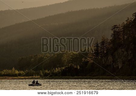 Two Fishermen In A Boat In The Lake Against The Backdrop Of The Forest And Mountains