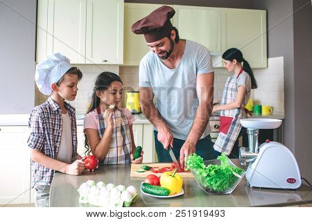 Positive Dad Cuts Tomato With Knife. Girl Holds Cucumber And Eats It. Boy Has Tomato In Hands And Lo
