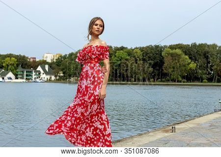 Portrait Of A Beautiful Young Brown-haired Girl With Long Hair In A Red Dress On A Lake Background.