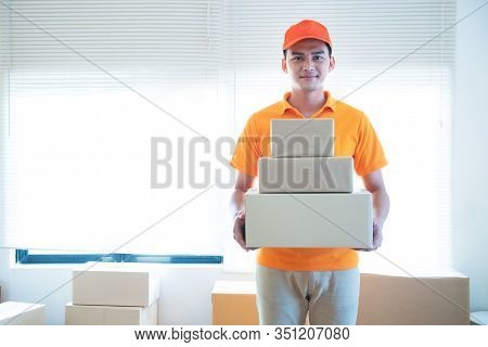 Smiling Young Delivery Asian Man In Uniform Holding And Carrying Stack Of Cardboard Postel In Office