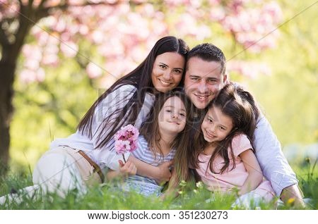Young Parents With Small Daugthers Sitting Outside In Spring Nature.