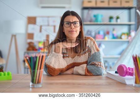 Young beautiful teacher woman wearing sweater and glasses sitting on desk at kindergarten cutting throat with hand as knife, threaten aggression with furious violence