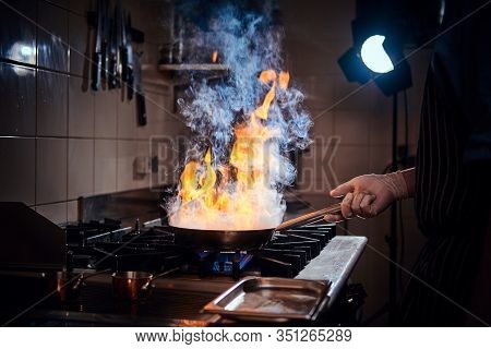 Professional Chef Wearing Gloves And Apron Cooking Stir-fry Flambe On A Pan With Open Fire In A Dark