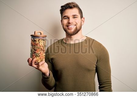 Young blond man with beard and blue eyes holding bottle of Italian dry pasta with a happy face standing and smiling with a confident smile showing teeth