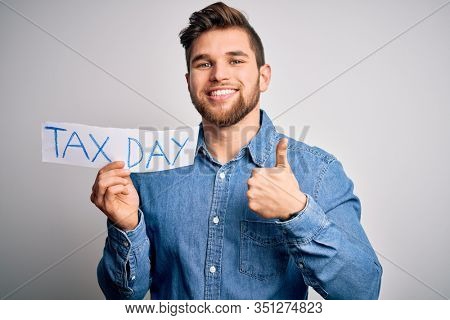 Young blond man with beard and blue eyes holding paper with tax day message happy with big smile doing ok sign, thumb up with fingers, excellent sign