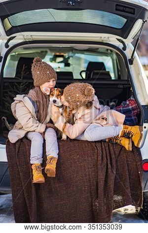 Two Sisters In Winter Clothes Sitting In The Trunk Of A Car With Dog And Play. It Is Winter And Snow