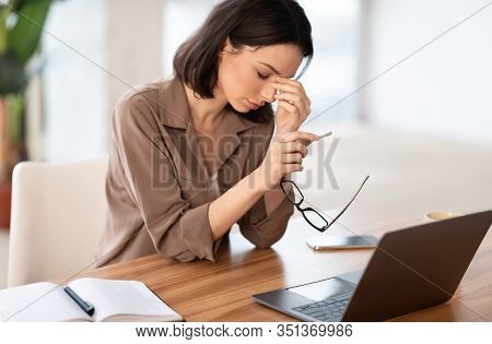 Eyes Fatigue. Tired Girl In Brown Shirt Massaging Her Nosebridge Holding Glasses Sitting At Desk Wit
