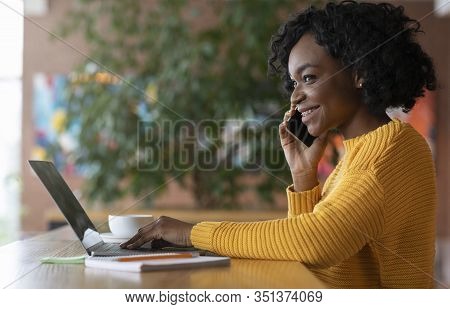 Smiling Young Black Woman Talking To Clients By Phone, Working Online In Cafe With Laptop, Copy Spac