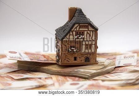A Ceramic House Stands On A Pile Of Five Thousand Ruble Bills. Symbol Of Success, Improvement Of Liv