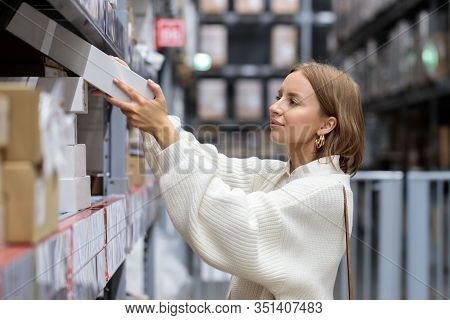 Female Customer In White Sweater Picks Up Goods In Self-service Warehouse, Side View. Shopping Conce