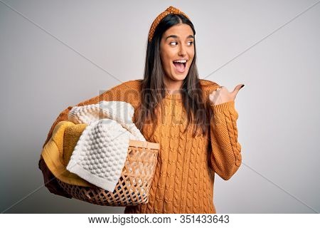 Young beautiful brunette woman doing housework chores holding wicker basket with clothes pointing and showing with thumb up to the side with happy face smiling