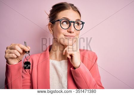 Young beautiful redhead house agent woman holding home key over pink bakcground serious face thinking about question, very confused idea