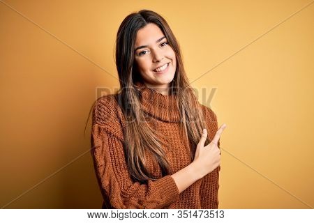Young beautiful girl wearing casual sweater standing over isolated yellow background cheerful with a smile of face pointing with hand and finger up to the side with happy and natural expression