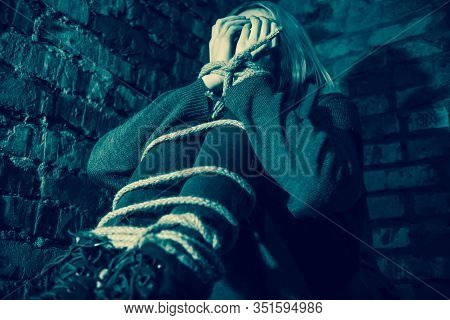 Blonde Girl With Bound Hands And Feet Is Sitting In The Basement, Covering Her Face With Her Hands A