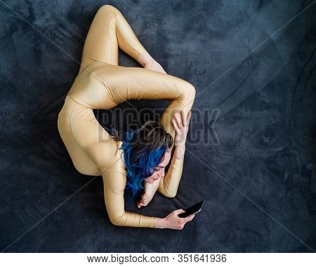 A Female Acrobat Lies On A Gray Bedspread In A Curved Pose And Uses A Mobile. A Woman Contortionist 