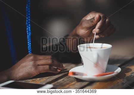 Close-up View Of Hands Of A Black Young Female Stirring Sugar In A Cup Of Coffee With A Spoon In An 