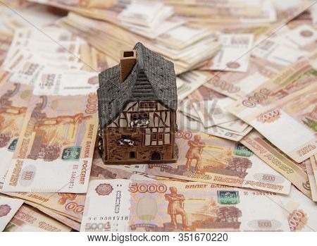 A Ceramic House Stands On A Pile Of Five Thousand Ruble Bills. Symbol Of Success, Improvement Of Liv